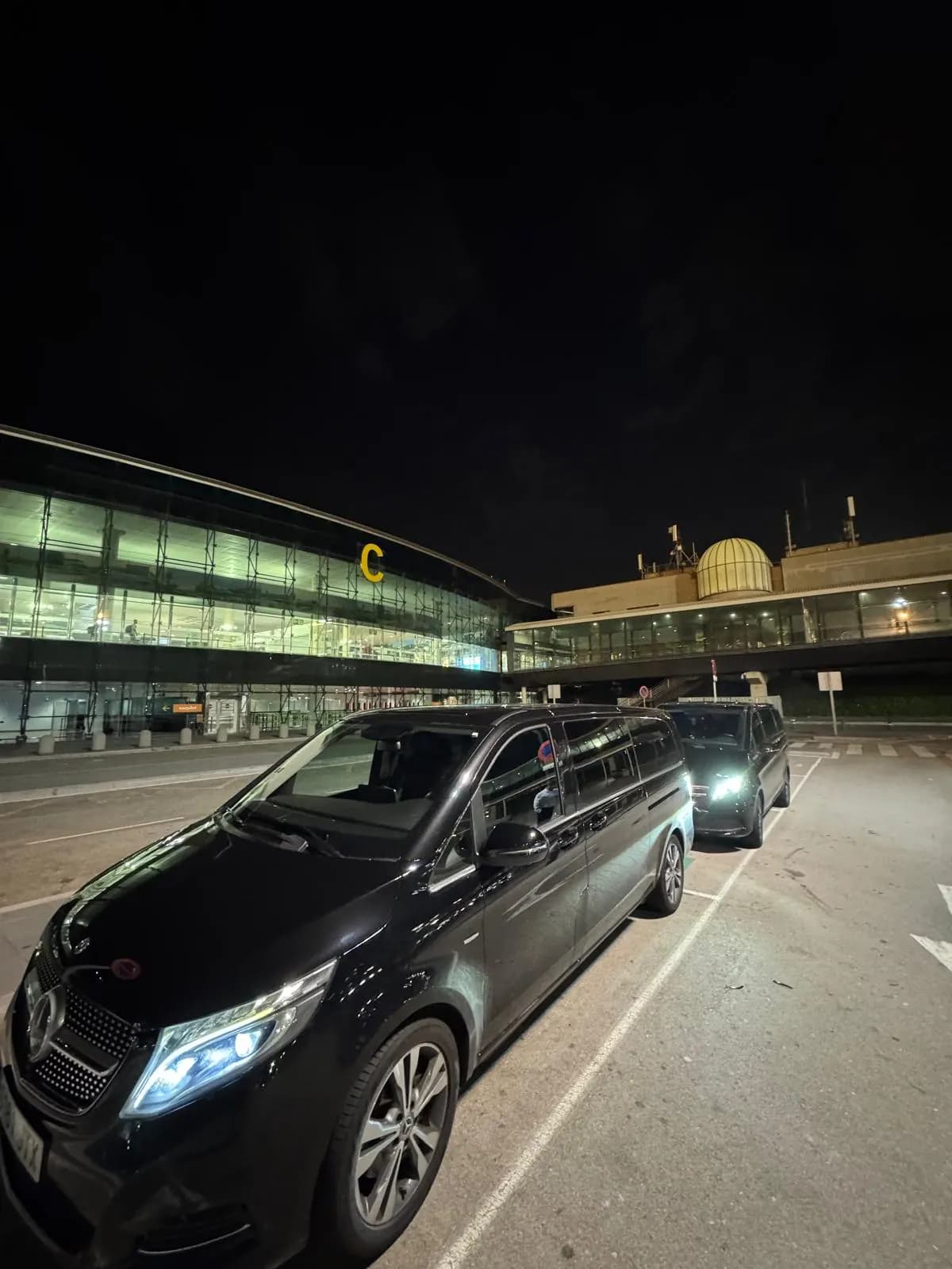 QuickPickups Mercedes V-Class with headlights on outside Barcelona Airport Terminal 1 at night